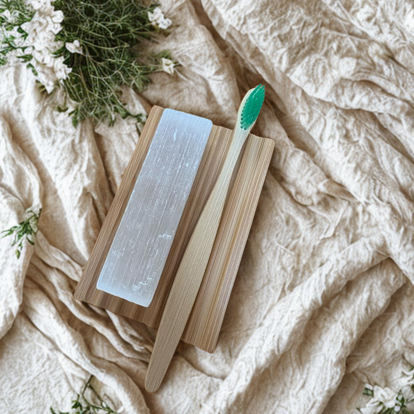 Bamboo toothbrush with a green bristle head on a wooden soap dish with selenite rod, placed on a textured fabric surface with floral elements.