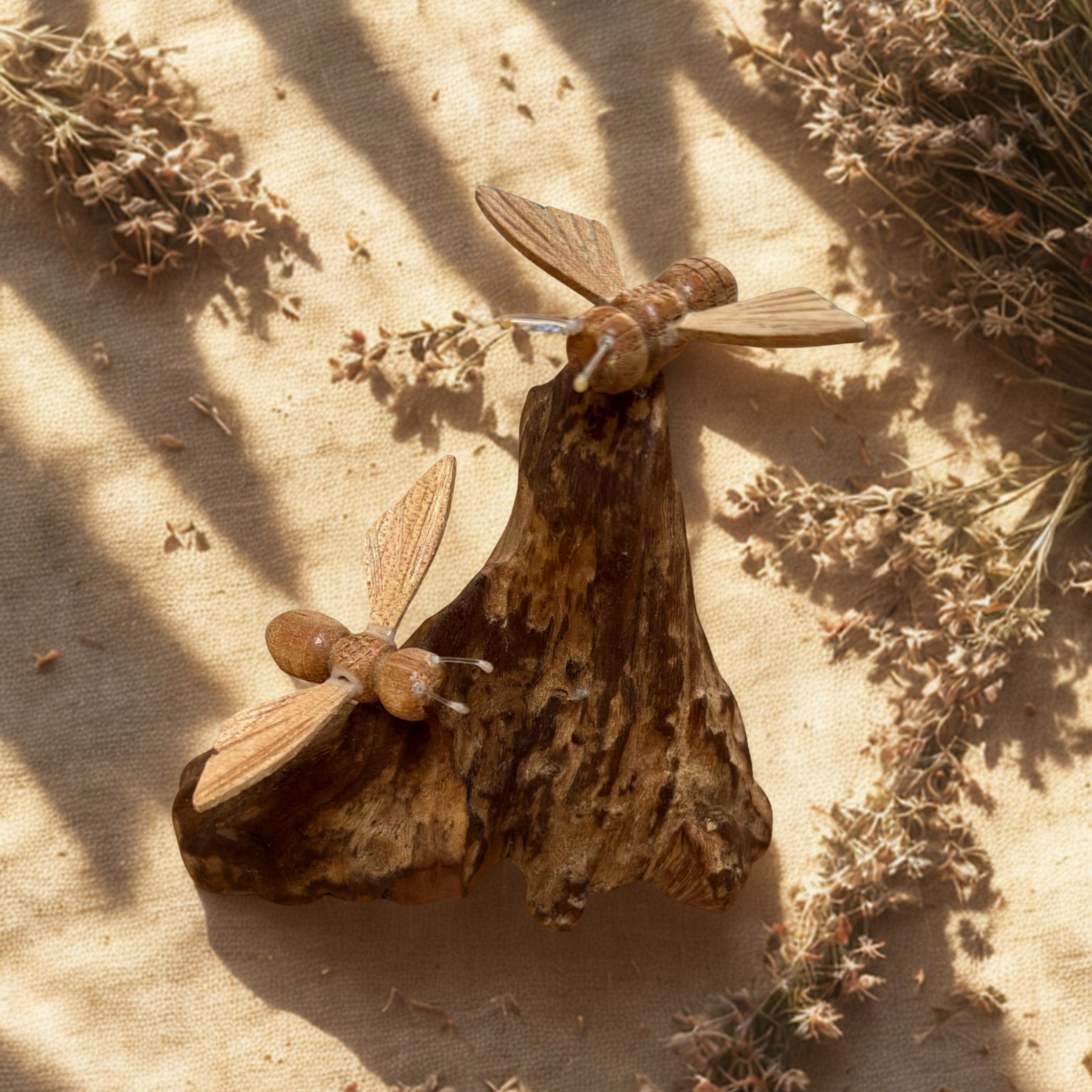 Two wooden bees on driftwood on a textured beige surface with dried plants.