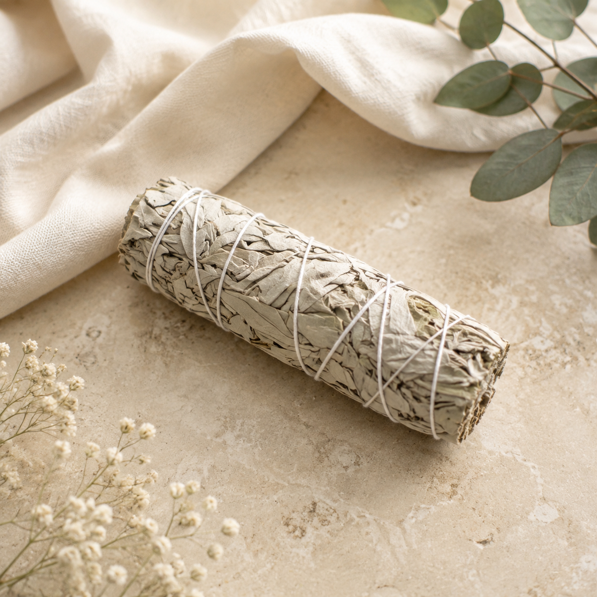 A dried sage smudge bundle wrapped in white string, styled on a textured beige stone surface with a cream linen cloth, green eucalyptus leaves, and small white baby's breath flowers.