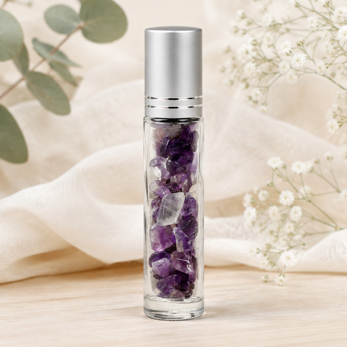 A clear glass roller bottle filled with amethyst crystal chips, topped with a brushed silver cap, set on a light wood surface with soft cream fabric and white flowers in the background.