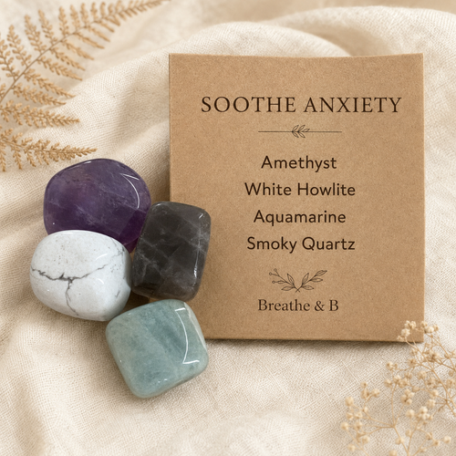 Four polished tumblel stones, Amethyst, White Howlite, Aquamarine, Smoky Quartz  are arranged beside a kraft paper card that reads “Soothe Anxiety” with stone names and “Breathe & B·.” The scene is styled on beige fabric with dried botanicals.