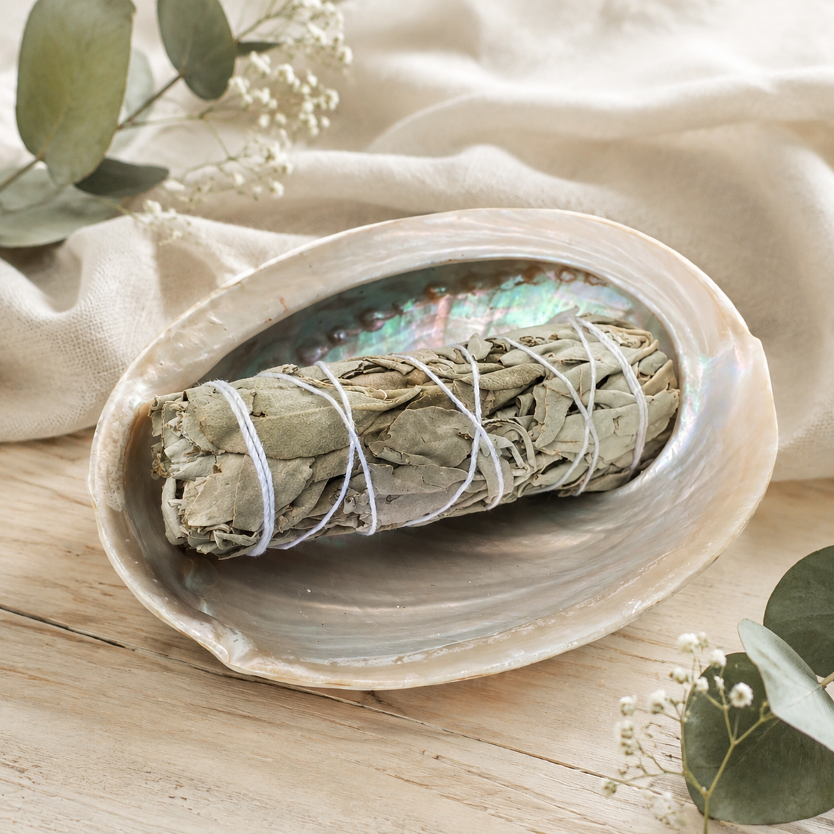A sage smudge bundle wrapped in white string rests inside an abalone shell dish on a light wood surface, surrounded by draped beige fabric, eucalyptus leaves, and small white flowers.