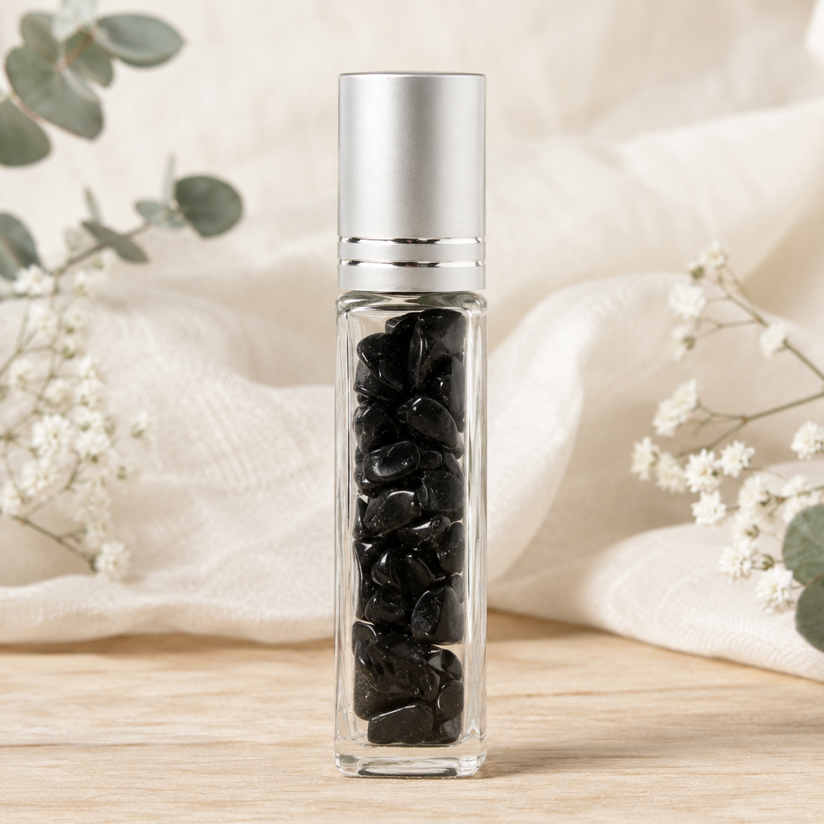 A clear glass roller bottle filled with black Obsidian Crystal Chips topped with a metallic silver cap, standing on a light wood surface with soft white fabric and small white flowers in the background.