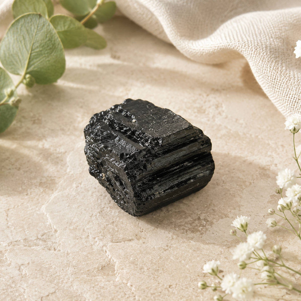 A black tourmaline cluster, layered texture, displayed on a textured beige stone surface with a draped cream linen cloth and small white flowers.