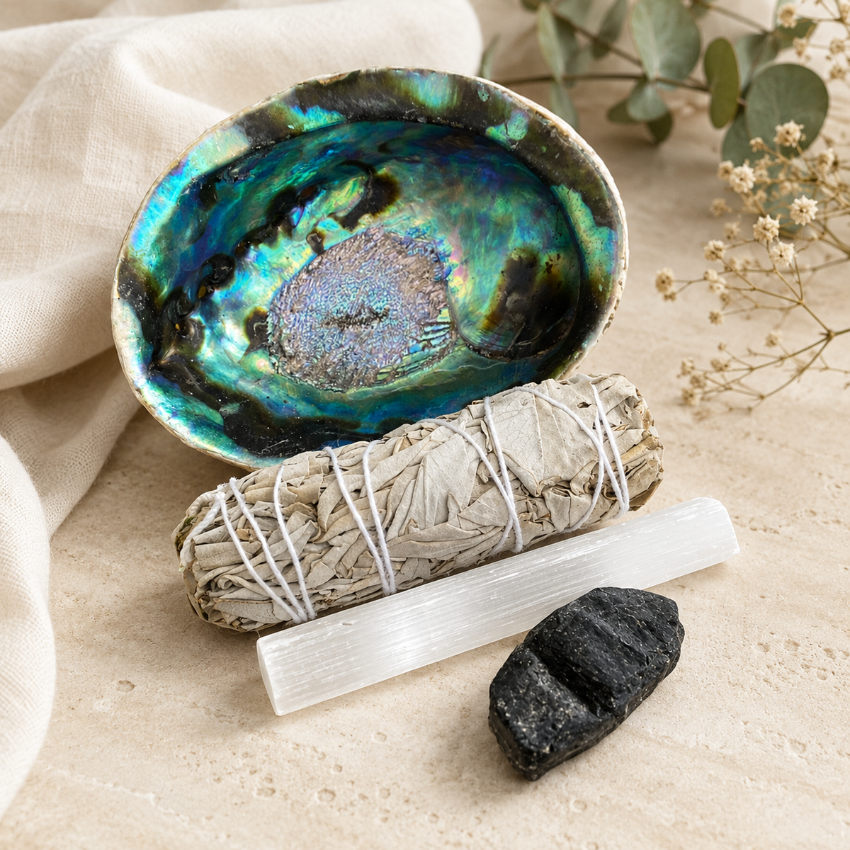 A natural abalone shell dish with a shimmering blue, green, and purple interior, styled with a sage smudge bundle, a clear quartz wand, and a black tourmaline cluster on a beige fabric surface.