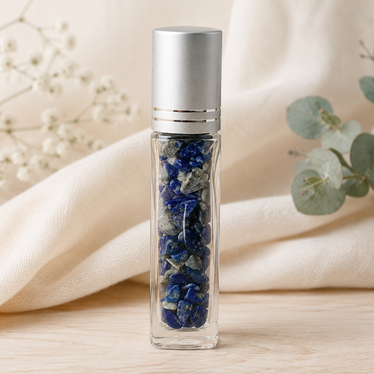 A clear glass roller bottle filled with lapis lazuli crystal chips, topped with a brushed silver cap, standing on a light wood surface with soft cream fabric and greenery in the background.