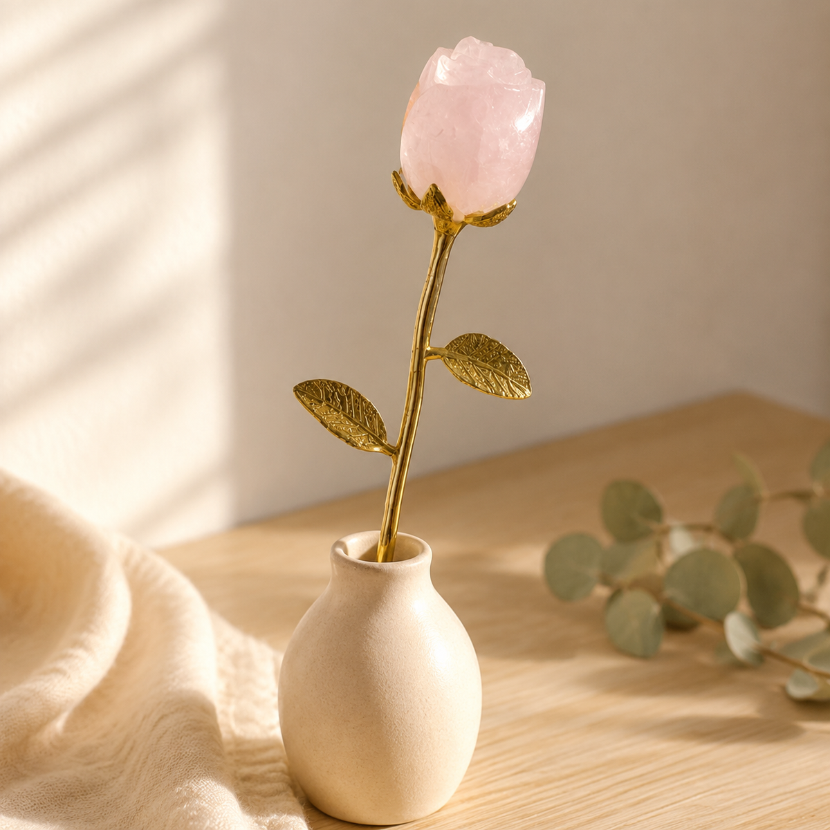 Rose quartz crystal flower with a gold metal stem and leaves displayed in a small cream vase on a softly lit wooden table.