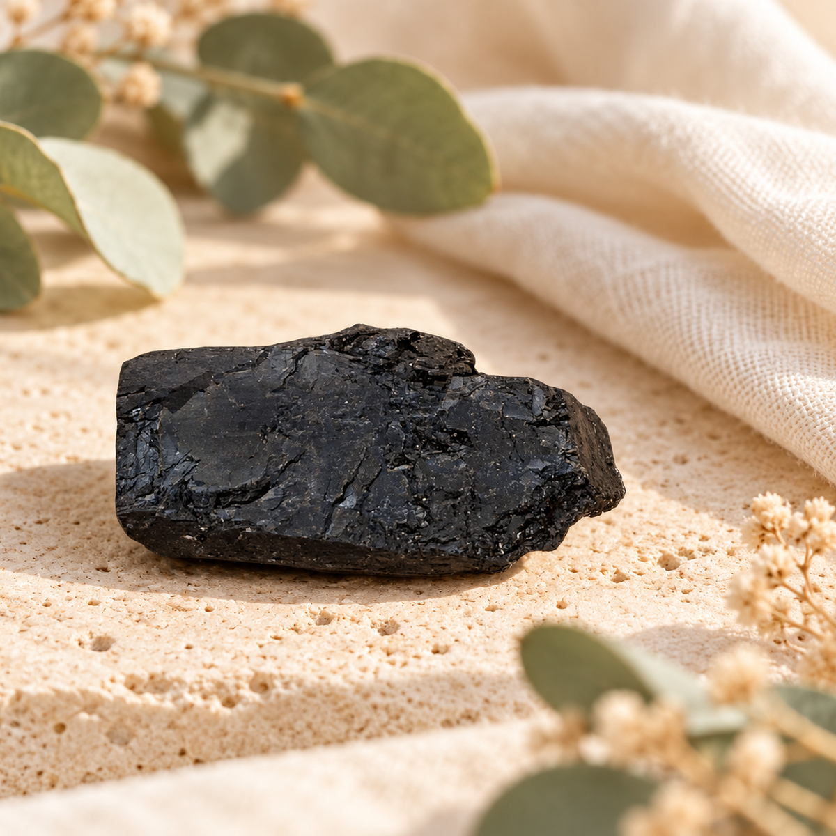 Chunk of raw black tourmaline with natural ridged texture, photographed on pale travertine with ivory linen and dried botanicals.
