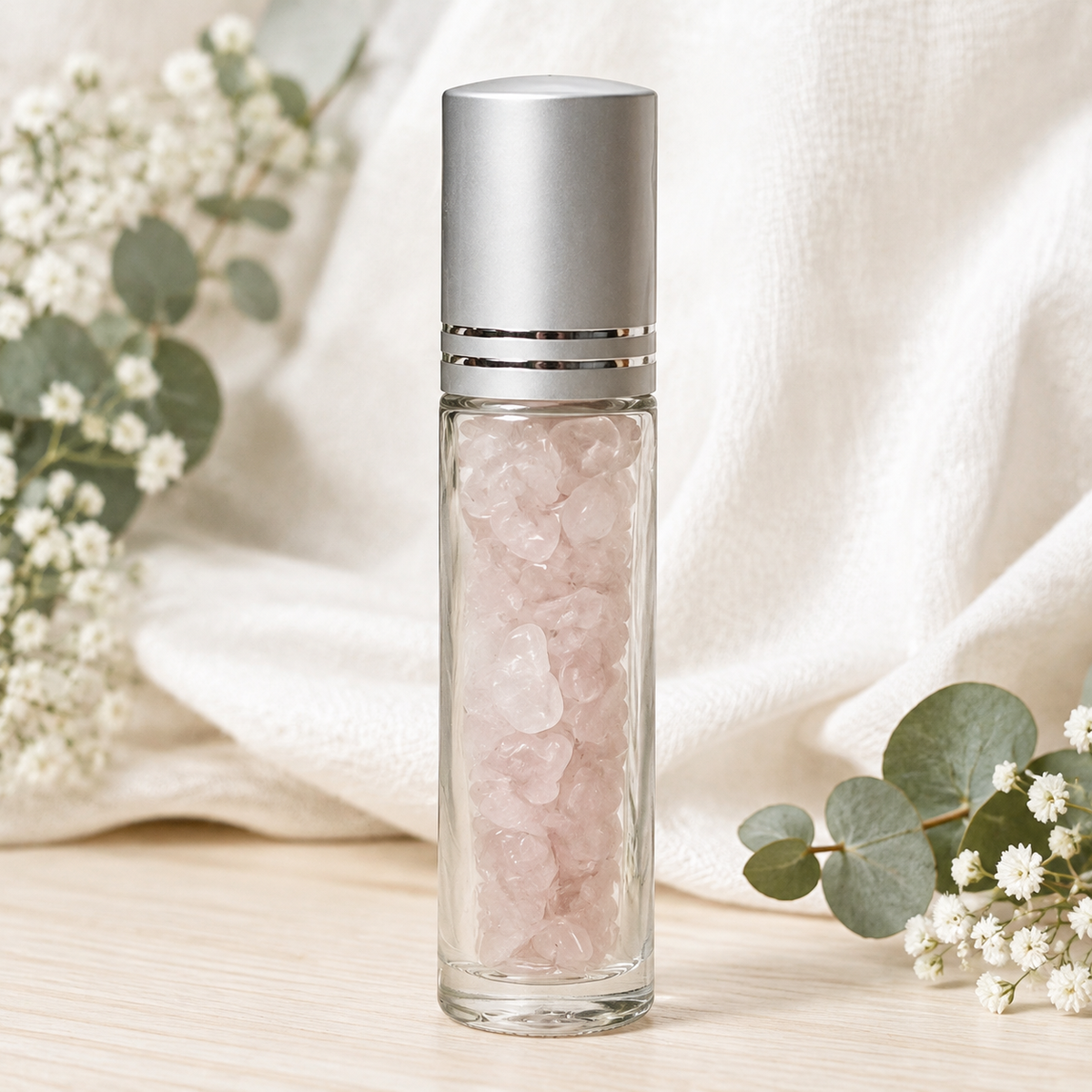A clear glass roller bottle filled with pale pink rose quartz crystals, topped with a brushed silver cap, set on a light wood surface with soft white fabric and eucalyptus flowers in the background.