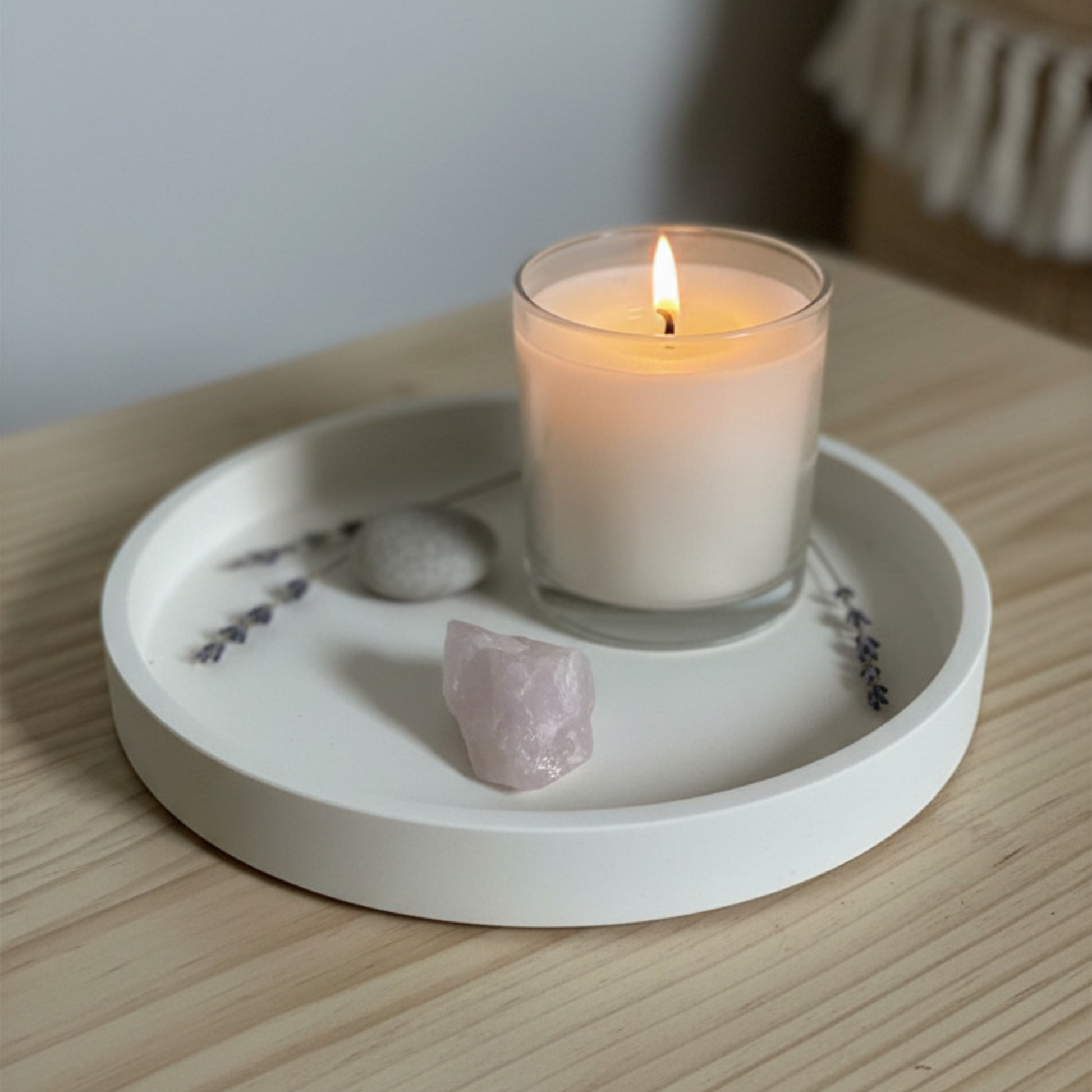 Candle in a glass holder on a white ceramic tray with lavender and a crystal on a wooden surface.