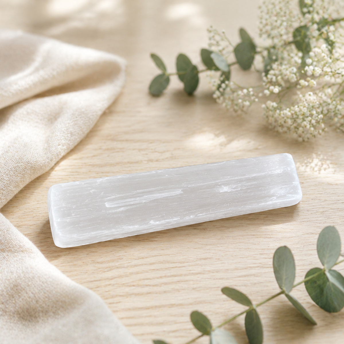 A rectangular white selenite crystal stone with natural striations, displayed on a light wood surface next to a beige fabric cloth and eucalyptus sprigs.