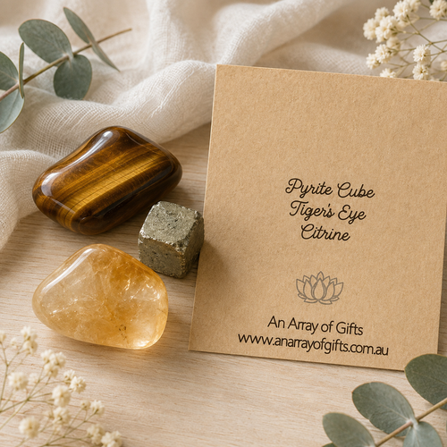 A gift set featuring a polished Tiger's Eye cube, a raw pyrite cube, and a Citrine stone arranged on a light wood surface with a beige fabric backdrop and a kraft paper card reading 'Pyrite Cube Tiger's Eye Citrine' and 'An Array of Gifts'.