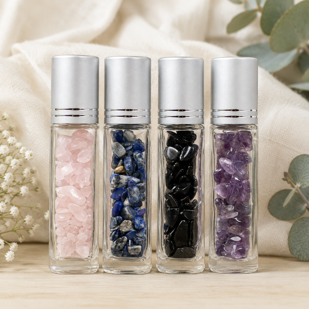Four clear glass roller bottles with silver caps, filled with different crystal stones in pink, blue, black, and purple, arranged on a light wood surface with a beige fabric backdrop and eucalyptus leaves.