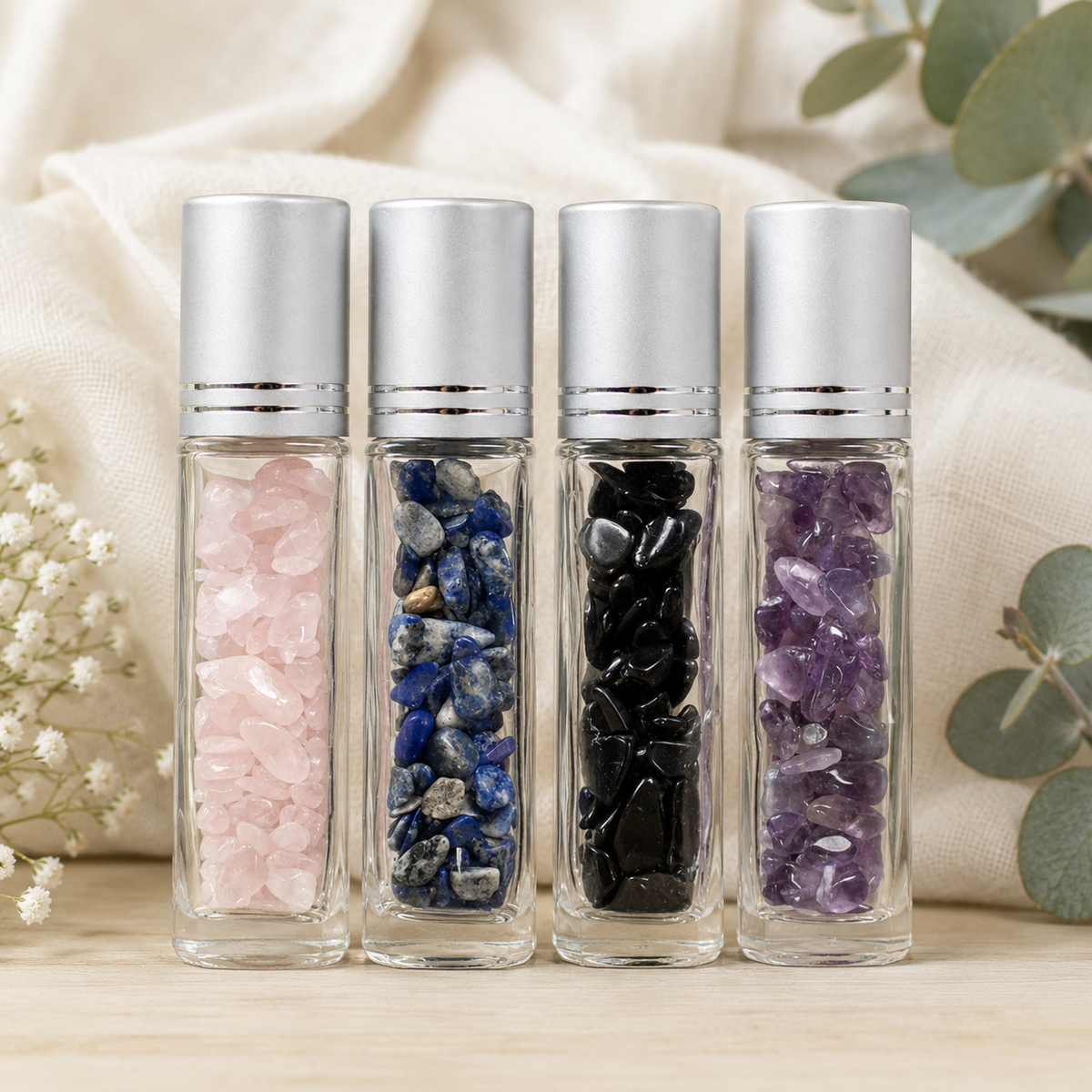 Four clear glass roller bottles with silver caps, filled with different crystal stones in pink, blue, black, and purple, arranged on a light wood surface with a beige fabric backdrop and eucalyptus leaves.