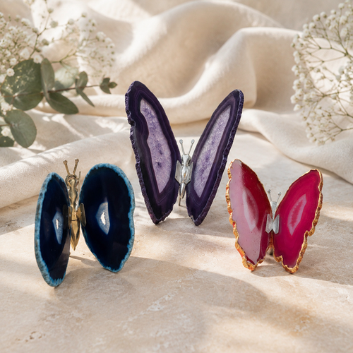 Pink, blue and purple agate butterfly ornaments displayed together on a warm neutral surface with ivory fabric and delicate greenery.