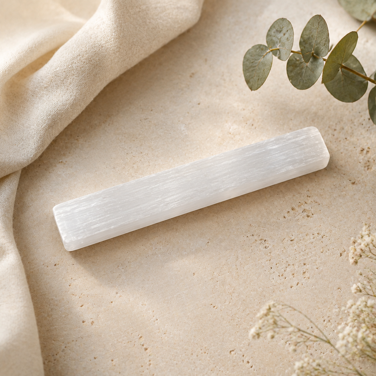 A white selenite wand resting on a textured beige stone surface, styled with a draped cream fabric, eucalyptus leaves, and small white flowers.
