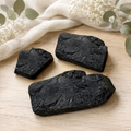 Three raw black tourmaline clusters with a textured, crystalline surface are arranged on a light wood surface with a cream linen cloth and dried white flowers in the background.