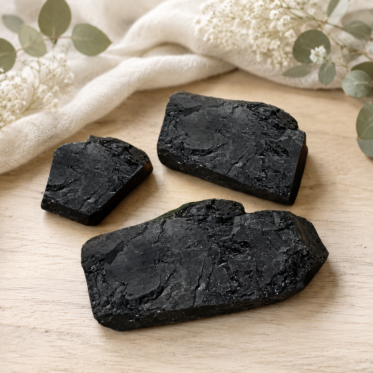 Three raw black tourmaline clusters with a textured, crystalline surface are arranged on a light wood surface with a cream linen cloth and dried white flowers in the background.