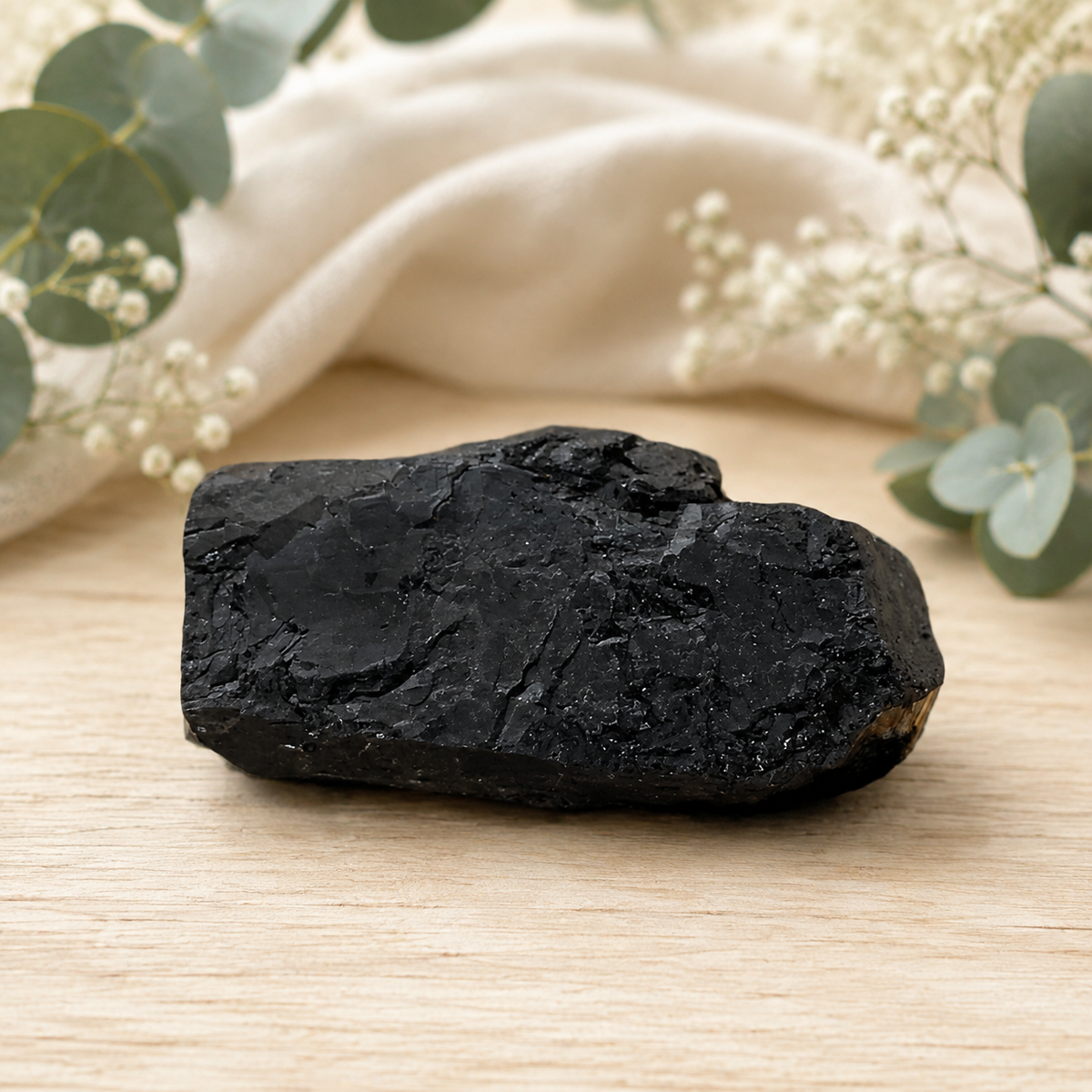 A raw black tourmaline cluster with a rough, textured surface, resting on a light wooden surface with a soft beige fabric and eucalyptus leaves in the background.