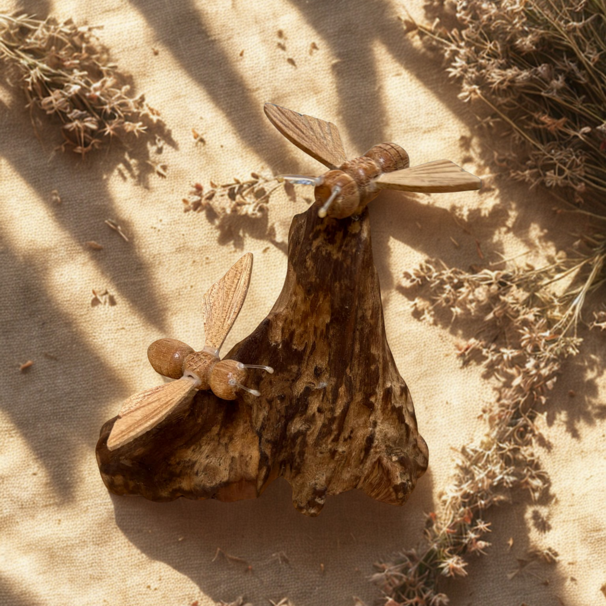 Two wooden bees on driftwood on a textured beige surface with dried plants.