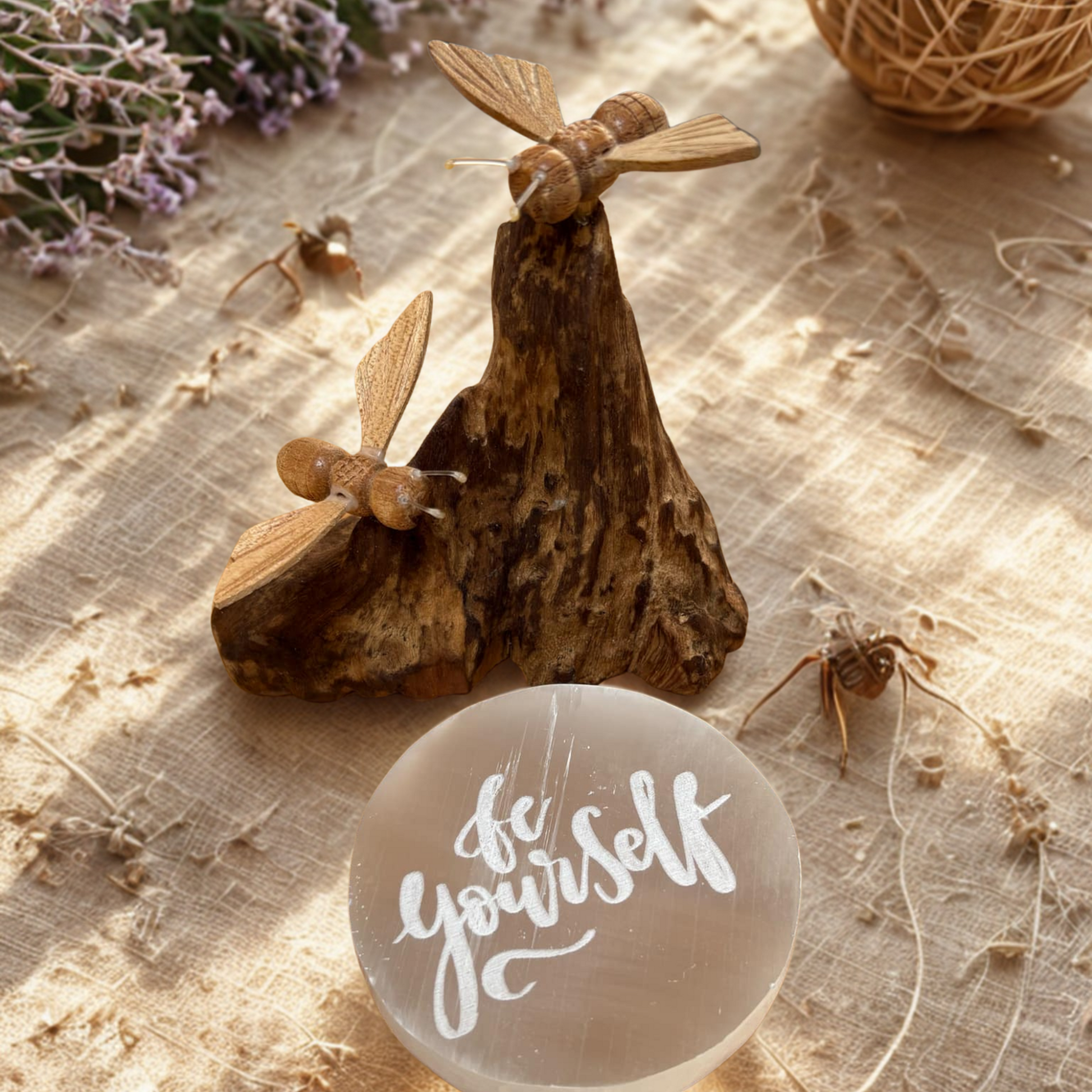 Wooden sculpture of bees on a log with a round stone featuring 'be yourself' text, on a textured surface.